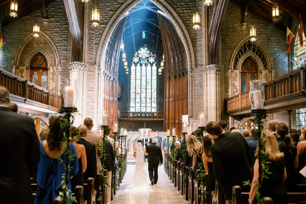This bride and her father approach the groom as the guests stand and an intricate stained glass window in the background is brightly lit by the sun.