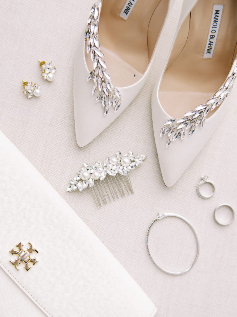 A flat lay of bridal Manolo Blahnik shoes, hair comb, earrings, bracelet, rings and purse featuring pearls and silver on pure white.