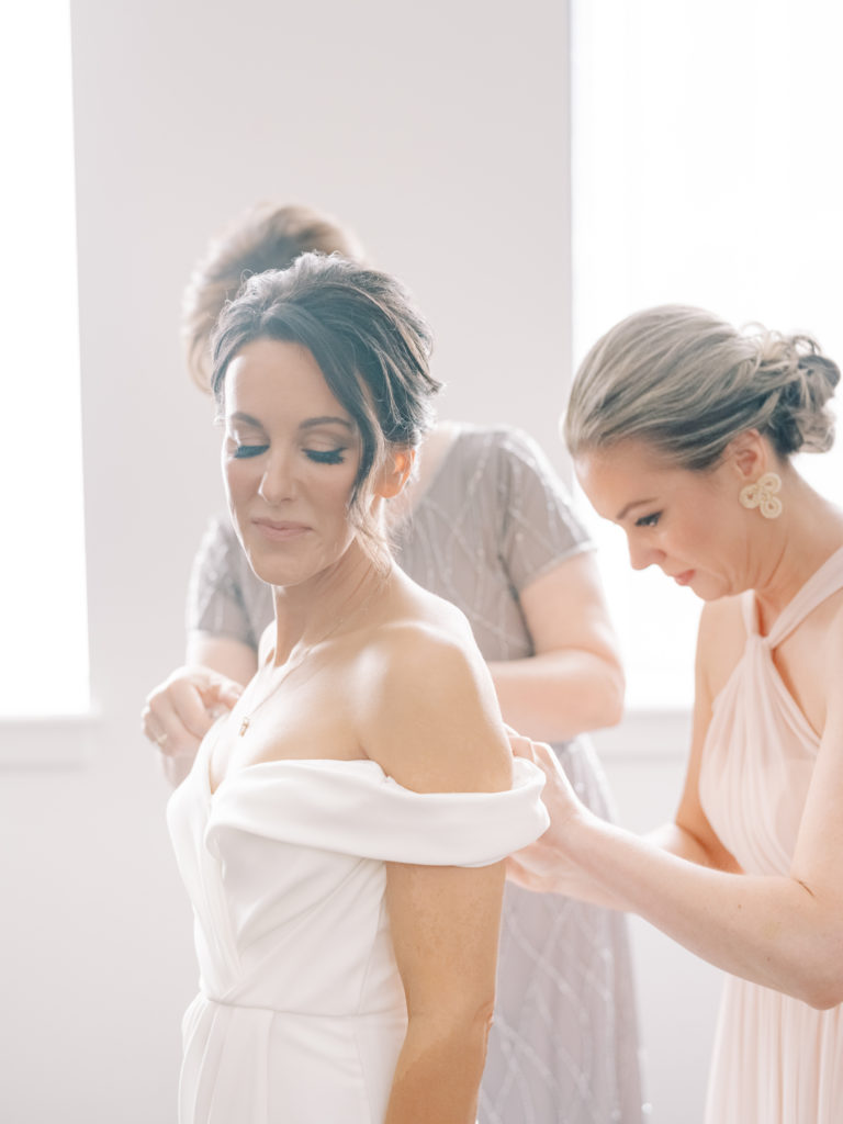 The bride glances softly down as her mother and maid of honor make some final adjustments to her dress by Anne Gregory.