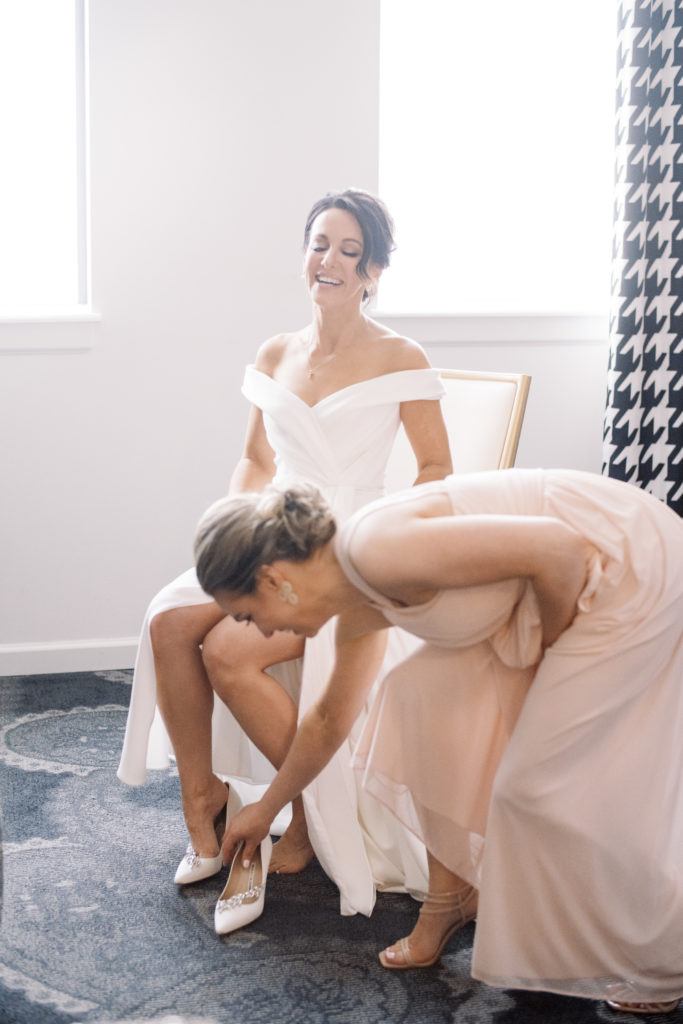 A bride's maid and the bride laugh as they work together to get the bride's Manolo Blahnik shoes on.